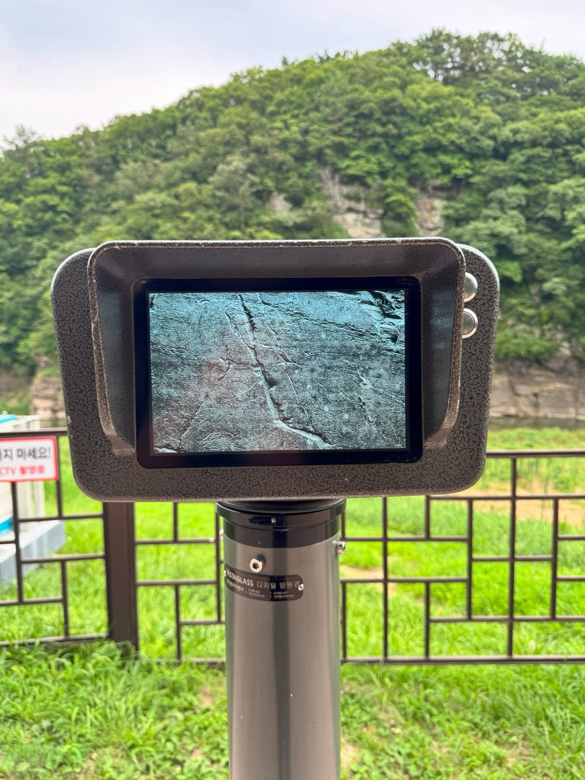 A viewing scope on a fence, its screen showing a magnified rock surface, with the cliff in the distance