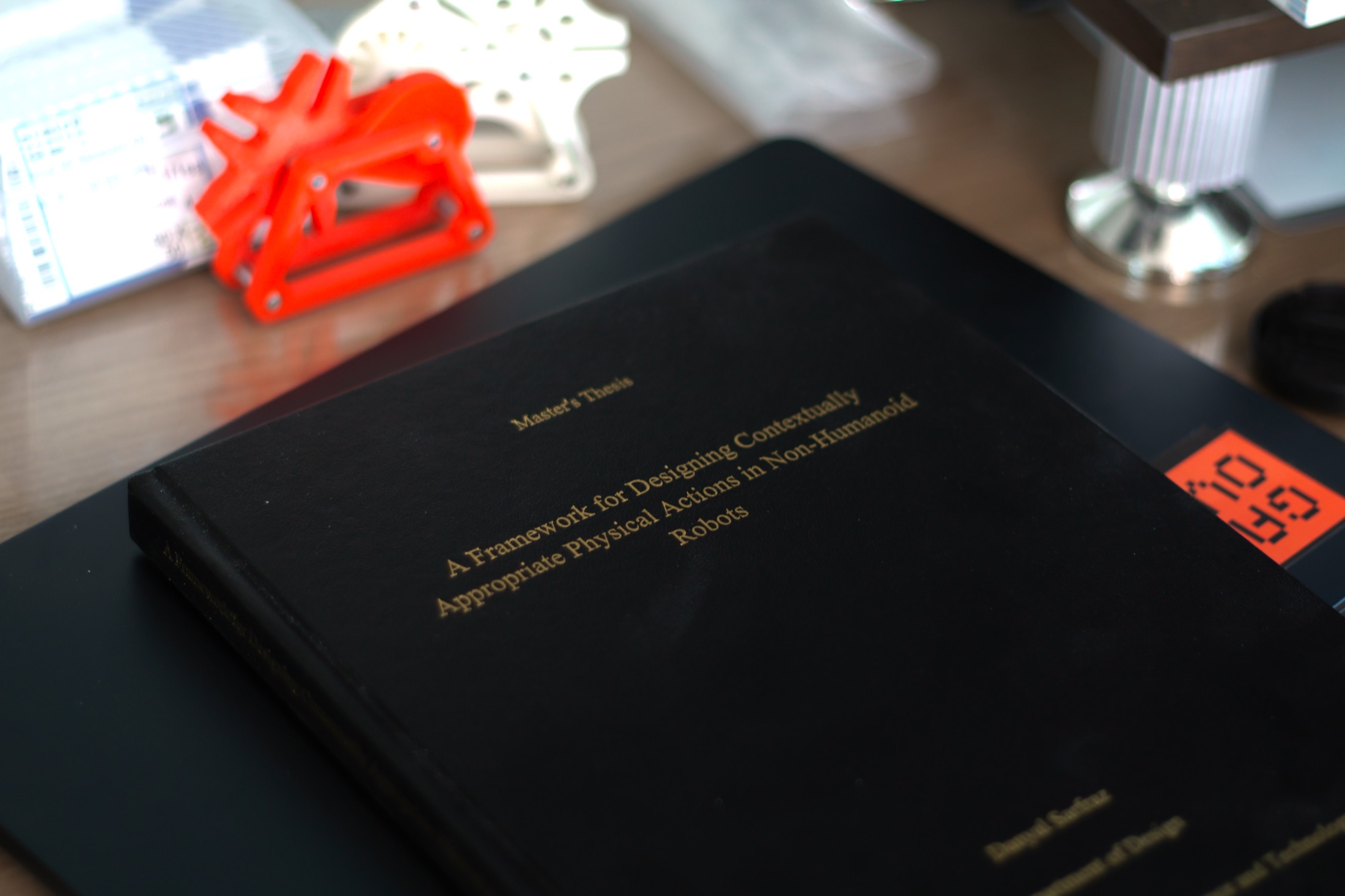 The bound thesis, a black hardcover titled A Framework for Designing Contextually Appropriate Physical Actions in Non-Humanoid Robots, next to an orange 3D-printed part