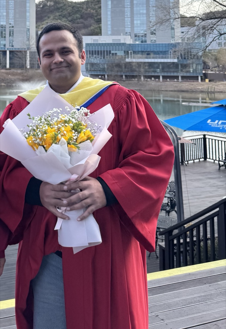 Danyal in red graduation robes, holding a bouquet of yellow flowers, with the UNIST campus behind